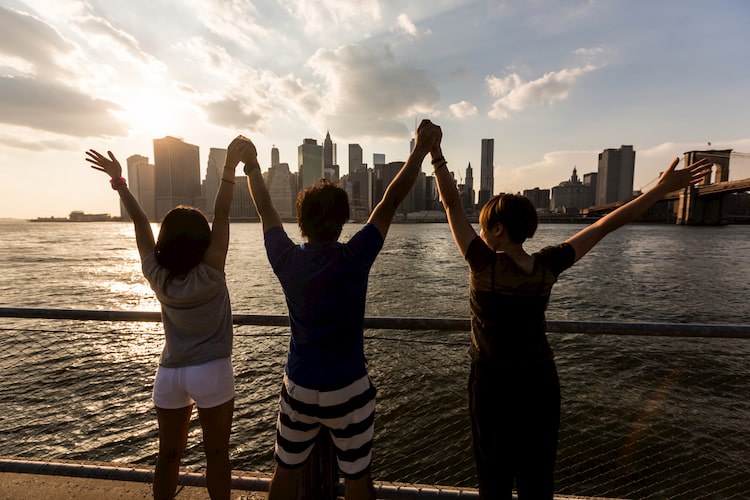 friends raise their arms as they look at the new york city skyline